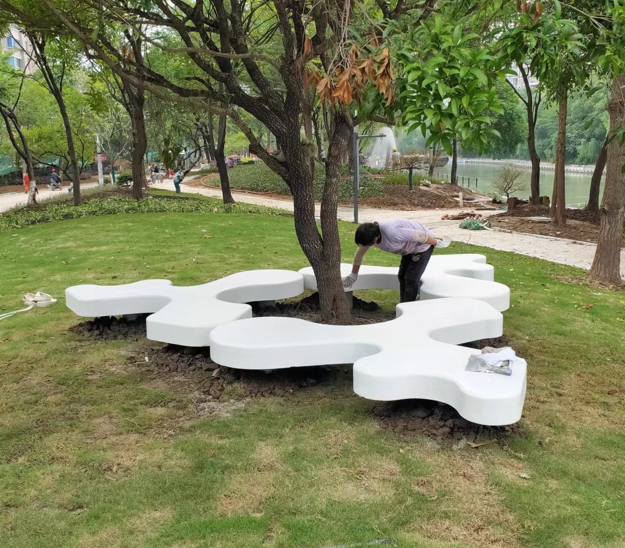 当混凝土跳出 “方正刻板” 的标签，这组异形坐凳成了公园的 “柔性焦点”—— 以流动的曲线包裹树基，让硬质设施与乔木生长空间自然咬合，打破了传统树池与坐具的边界感。 它的形态并非刻意的艺术炫技，而是以 “共生” 为核心逻辑：不规则的轮廓既适配树木的自然生长形态，也让坐区呈现出 “松散却聚拢” 的社交尺度 —— 既不会因过度紧凑带来压迫感，也能让休憩者在视觉联通中保持舒适距离。 白色的哑光质感弱化了混凝土的生冷，与草地、树冠的自然色调形成柔和反差，既是功能载体，也是场地里的 “视觉软装饰”。这种设计跳出了 “设施只是工具” 的惯性，让坐凳成为自然与空间的衔接媒介，把 “实用” 和 “场域美感” 揉成了公园里的日常风景。